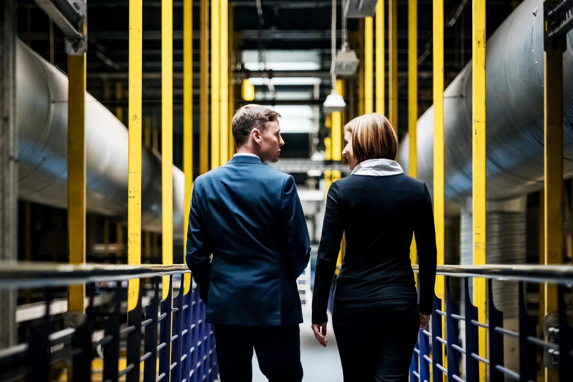 Eine Frau und ein Mann gehen durch eine Frabrik und unterhalten sich dabei.